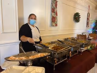A staff member wearing a face mask stands behind a buffet table with multiple trays of food in a dining area decorated with a Christmas wreath and holiday decorations.