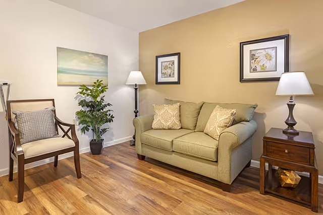 A cozy living room area with a green loveseat sofa adorned with patterned cushions, a wooden side table with a lamp, a wooden chair with a cushion, a potted plant, and two framed floral artworks on the wall. The room has wood flooring and soft lighting.
