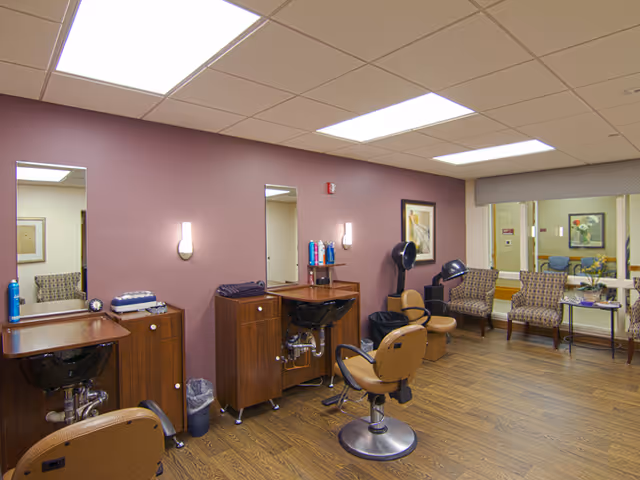 Interior of a senior living facility hair salon with two styling stations featuring mirrors, chairs, and sinks. There are hair care products on the counters and two hair dryers with chairs nearby. The room has wooden flooring, mauve walls, and a seating area with three patterned chairs and a small table near a window.