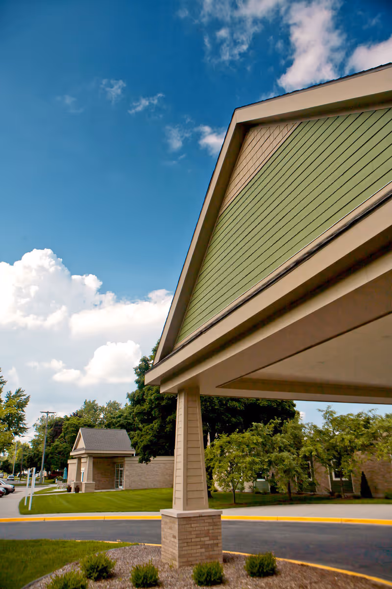 View of the exterior entrance canopy of The Cortland Wyoming facility with a clear blue sky and some clouds in the background. The canopy has a green and beige color scheme with brick pillars, and there are trees and a smaller building visible in the background.