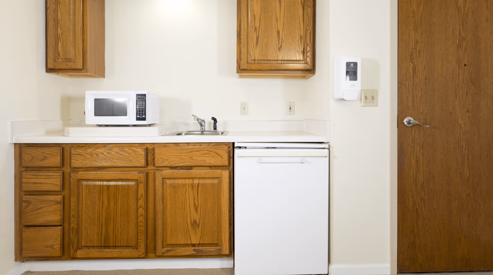Small kitchenette with wooden cabinets, a microwave on the counter, a sink and a compact refrigerator next to a wooden door.