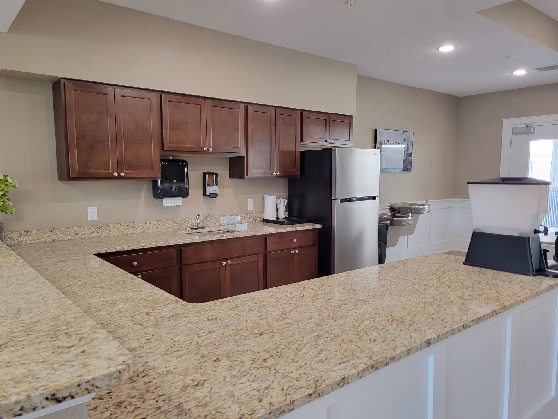 A clean kitchen area with granite countertops and dark wooden cabinets. There is a stainless steel refrigerator, a sink, a paper towel holder, a soap dispenser, and a water fountain mounted on the wall. The walls are beige, and there is a framed picture hanging near a door with a window.