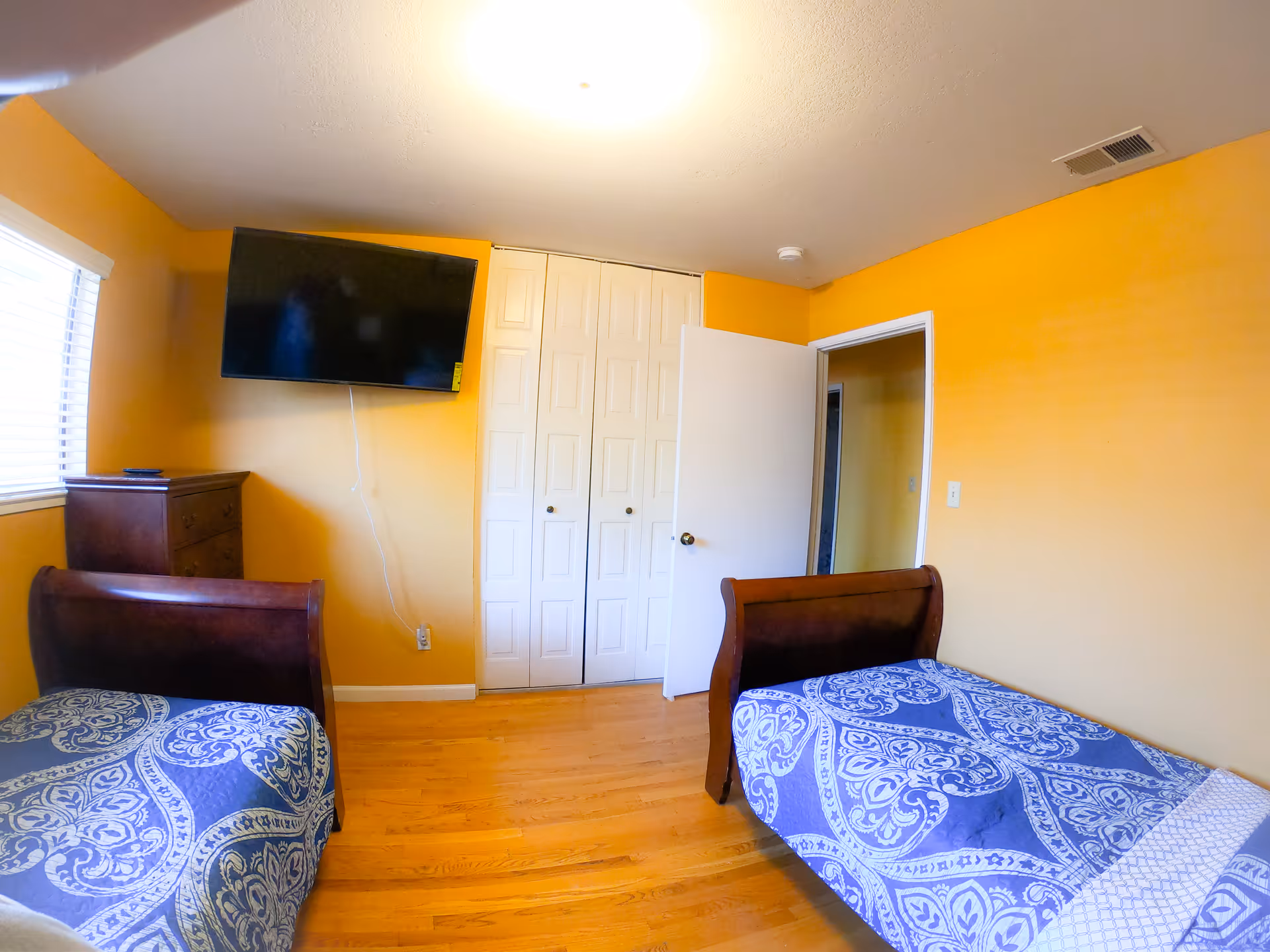 A bedroom with two single beds featuring blue and white patterned bedspreads. The walls are painted yellow, and there is a wooden dresser under a window with blinds. A flat-screen TV is mounted on the wall above the dresser. A white door is open, and a white closet with double doors is visible.