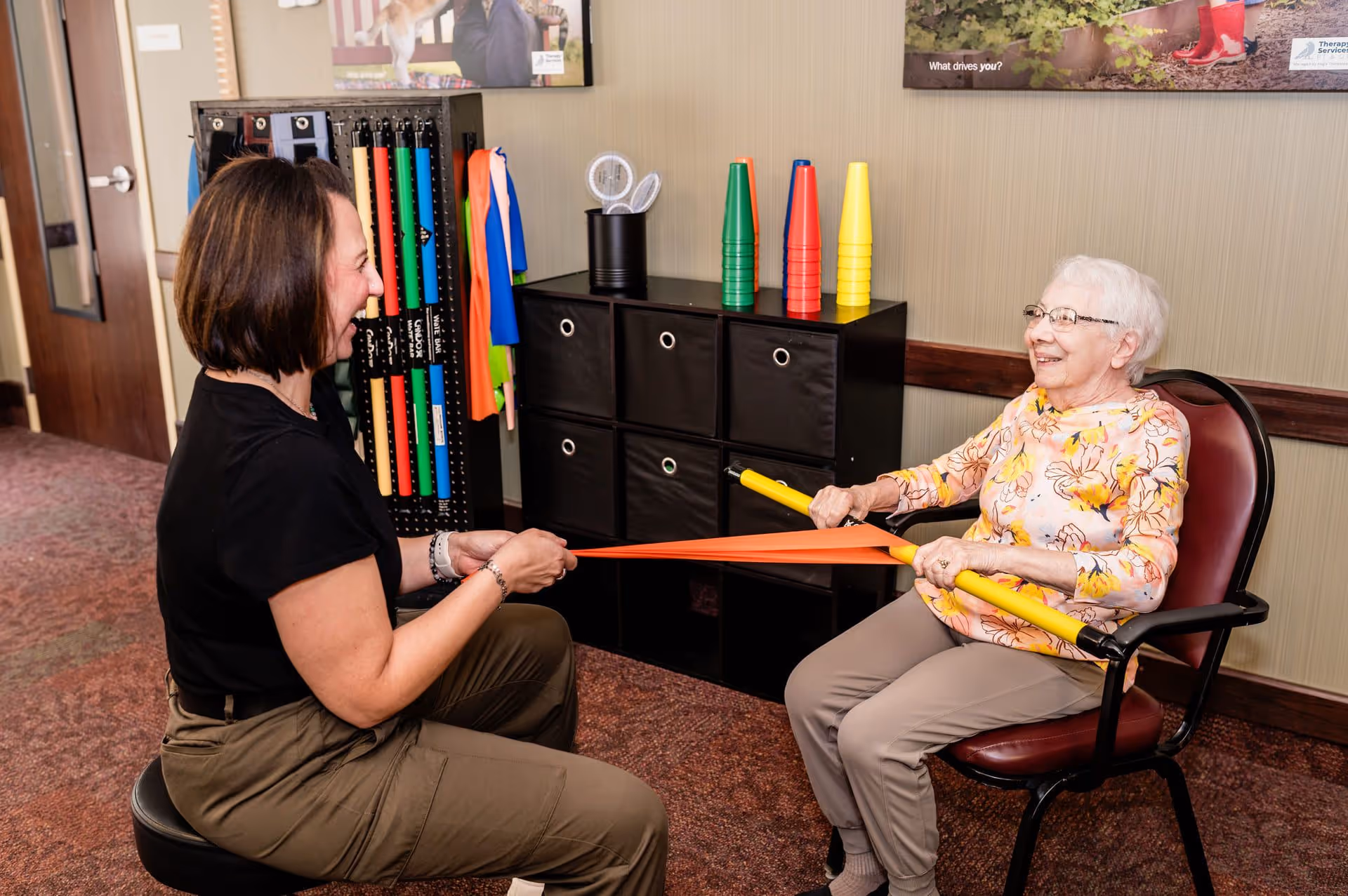 An elderly woman sitting in a chair and a younger woman sitting on a stool are engaging in a physical therapy exercise using a yellow bar and an orange resistance band in a room with storage cubes and colorful cones in the background.