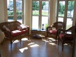 A bright sitting area with wicker furniture including a loveseat, two armchairs with red cushions, and a glass-top coffee table. Large windows and a glass door provide a view of a green outdoor space with houses in the background.