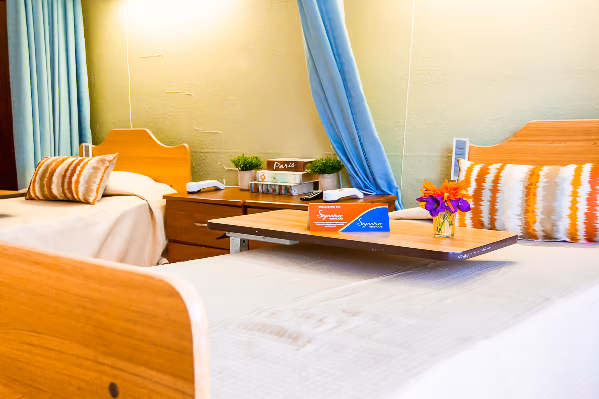A bright and cozy bedroom with two single wooden beds, each with patterned pillows. Between the beds is a wooden nightstand with potted plants, books, and remote controls. A blue curtain hangs in the background, and a wooden overbed table with a small vase of colorful flowers and a Signature HealthCARE welcome card is placed on one of the beds.