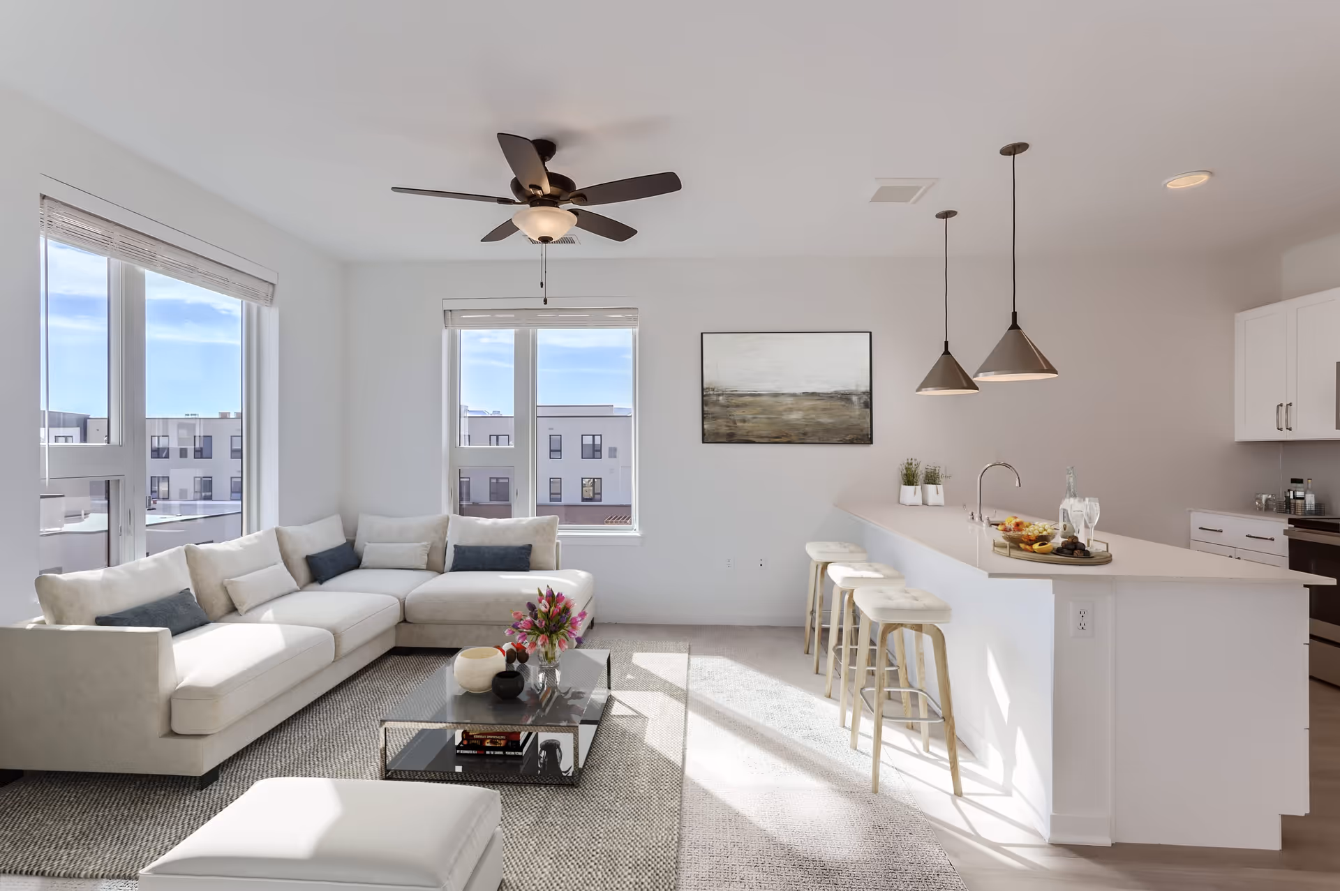 Bright and modern living room with a large white sectional sofa adorned with cushions, a glass coffee table with decorative items and flowers, a ceiling fan with light, and large windows letting in natural light. Adjacent to the living area is a white kitchen island with three bar stools, pendant lights hanging above, and a kitchen area with white cabinets and appliances.