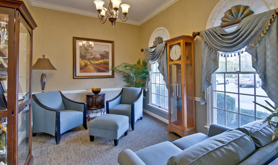 A cozy living room area with two blue armchairs, a matching ottoman, and a blue sofa. There is a wooden grandfather clock between two large windows with decorative blue curtains. A small wooden side table with a lamp and a bowl is placed between the armchairs. A framed landscape painting hangs on the beige wall, and a green potted plant is near the windows.