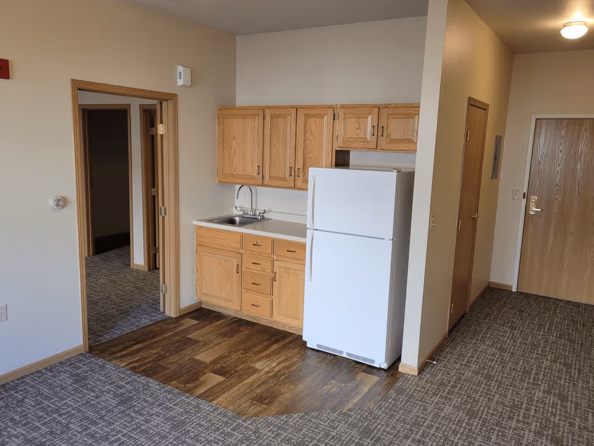 Interior view of a senior living facility unit featuring a small kitchen area with wooden cabinets, a white refrigerator, and a sink. The floor transitions from carpet to wood near the kitchen. There are two doorways visible, one leading to another room and the other to a hallway with a closed wooden door at the end.