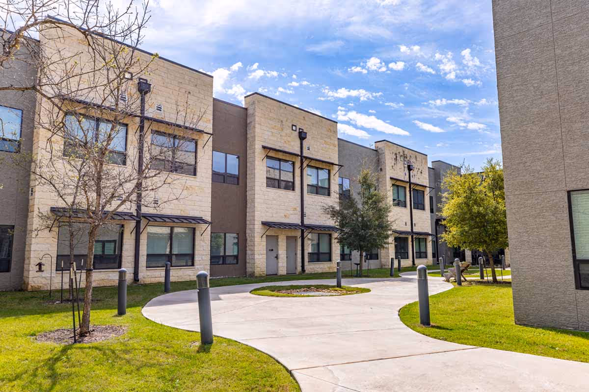 Exterior view of a modern senior living facility with beige and gray buildings, large windows, a curved concrete walkway, green grass, and small trees under a partly cloudy blue sky.