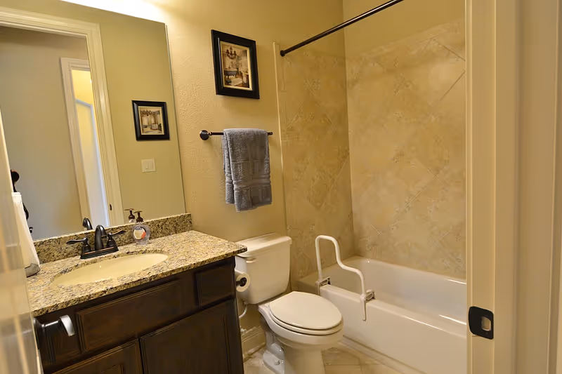 Well-lit bathroom with a granite vanity and sink, toilet, and a bathtub-shower with a safety grab bar and tiled walls.
