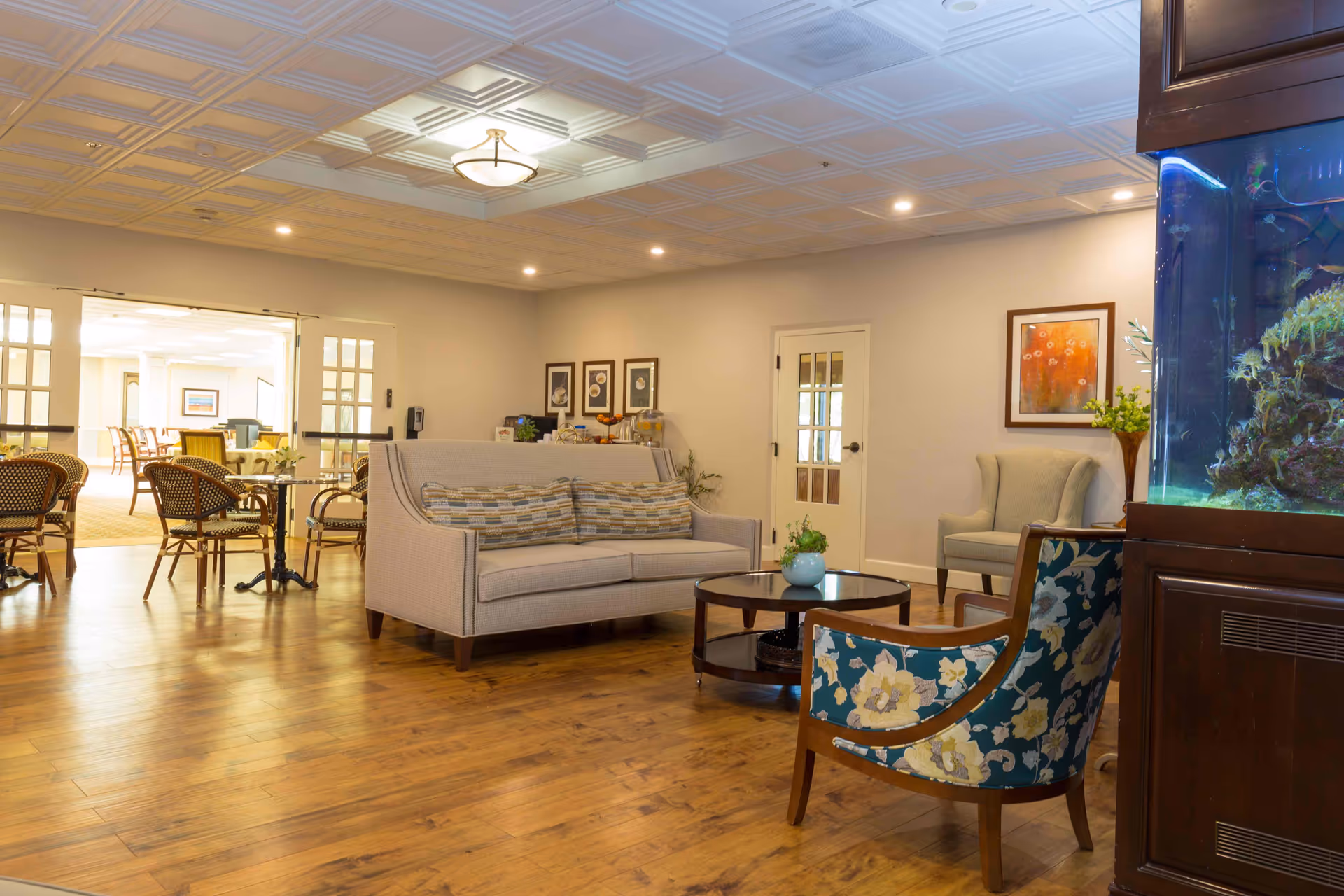 A cozy senior living facility common area with wooden flooring, a beige sofa with patterned cushions, a round wooden coffee table with a small plant, and two armchairs, one with floral upholstery. The room is well-lit with ceiling lights and has framed artwork on the walls. In the background, there is a dining area with tables and chairs visible through glass-paneled double doors. On the right side, there is a large aquarium integrated into a wooden cabinet.