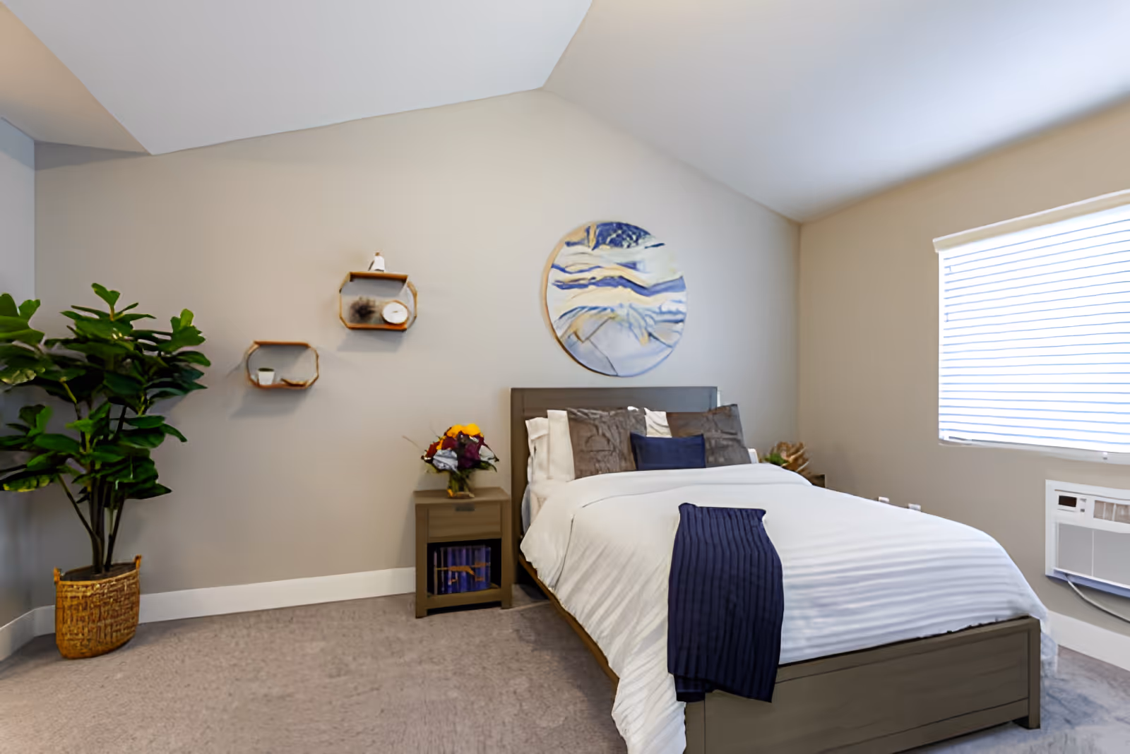 A neatly made bed with white bedding and a dark blue throw blanket in a bedroom with beige walls. Above the bed is a round abstract painting with blue and white tones. To the left of the bed is a small nightstand with a vase of flowers and two hexagonal wall shelves with decorative items. A large green potted plant is in the corner. A window with closed blinds and an air conditioning unit is on the right wall.