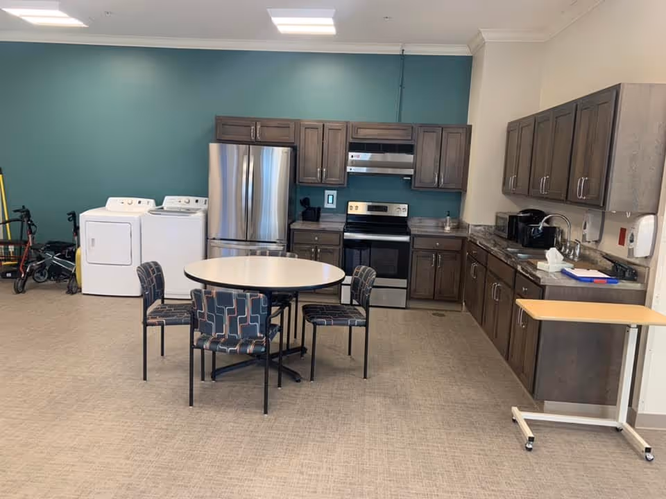 A kitchen and laundry area in a senior living facility with a round table and four patterned chairs in the center. The kitchen has wooden cabinets, a stainless steel refrigerator, stove, and a countertop with a sink and various appliances. To the left, there is a white washing machine and dryer. The walls are painted teal and white, and the floor is covered with beige carpet.