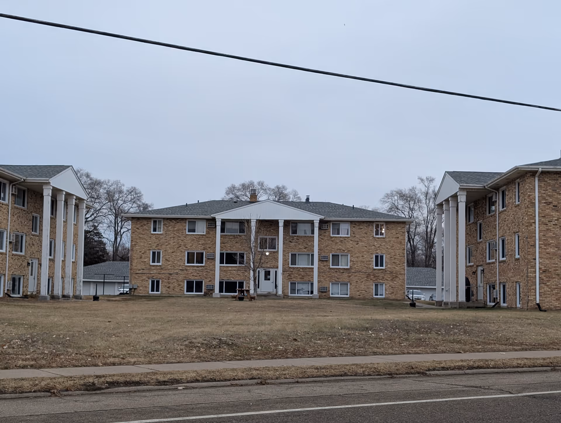 Exterior view of three connected brick apartment buildings with white columns on the front, set on a grassy area with a sidewalk and street in the foreground under an overcast sky.