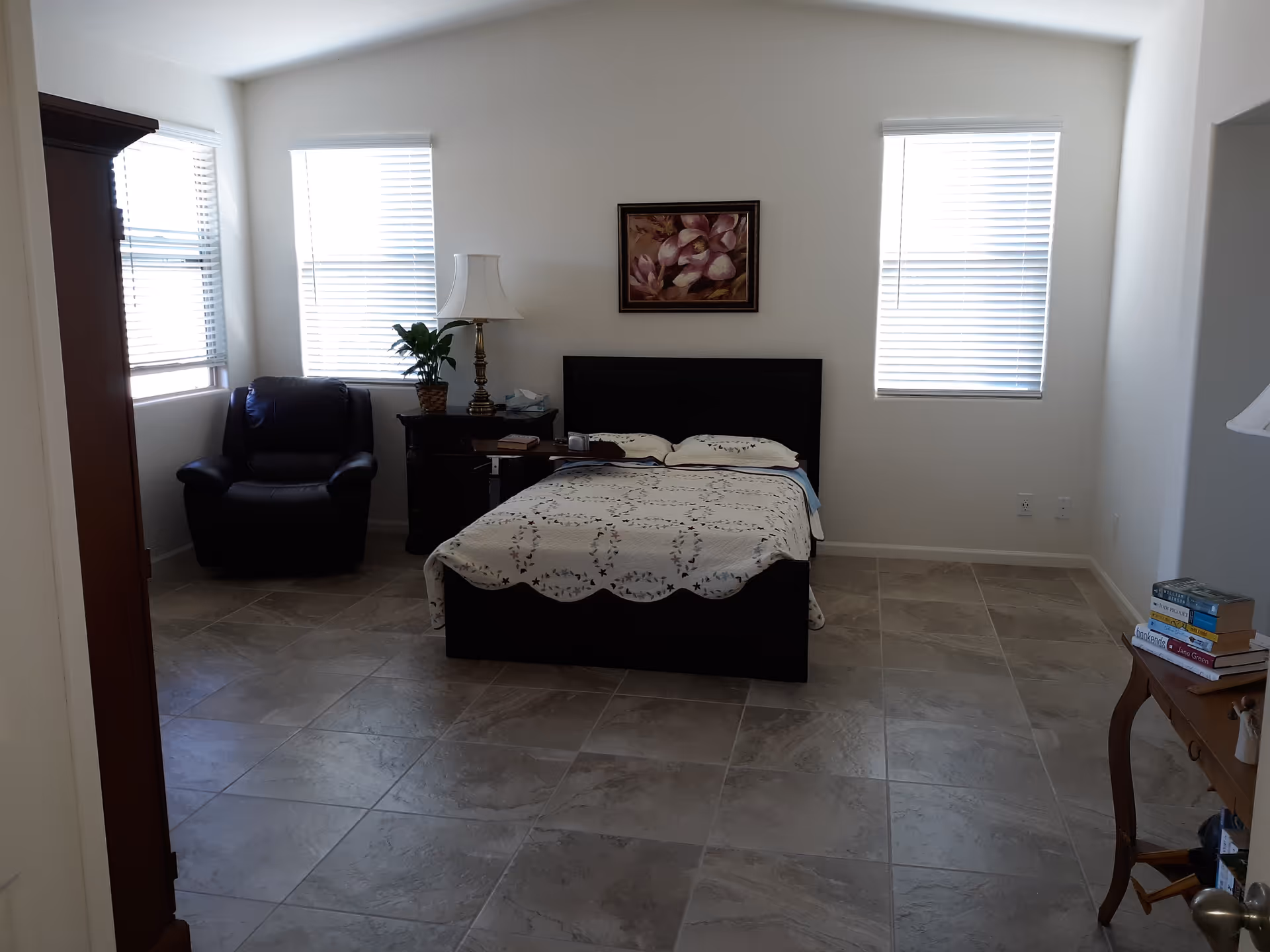 A bedroom with a bed covered in a white patterned bedspread, two pillows, and a dark wooden headboard. There are two windows with blinds letting in natural light. To the left of the bed is a dark armchair and a nightstand with a lamp and a plant. On the right side, there is a small wooden table with a stack of books. The floor is tiled with large square tiles, and a framed floral painting hangs on the wall above the bed.