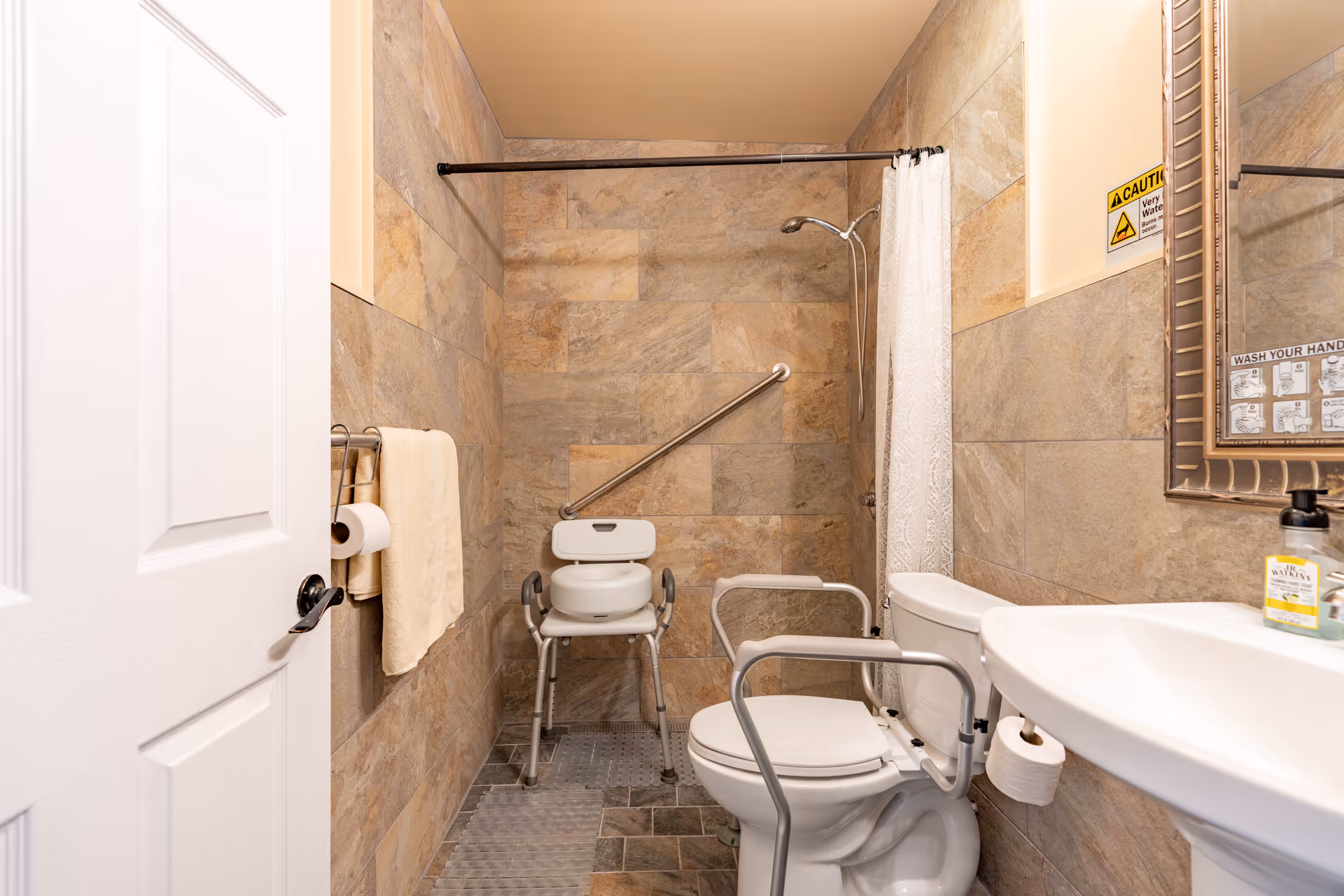 Accessible bathroom with beige tiled walls and floor, featuring a white toilet with safety rails, a white shower chair, a shower area with a curtain, a white sink with a soap dispenser, a towel rack with a towel and toilet paper, and safety warning signs on the wall.