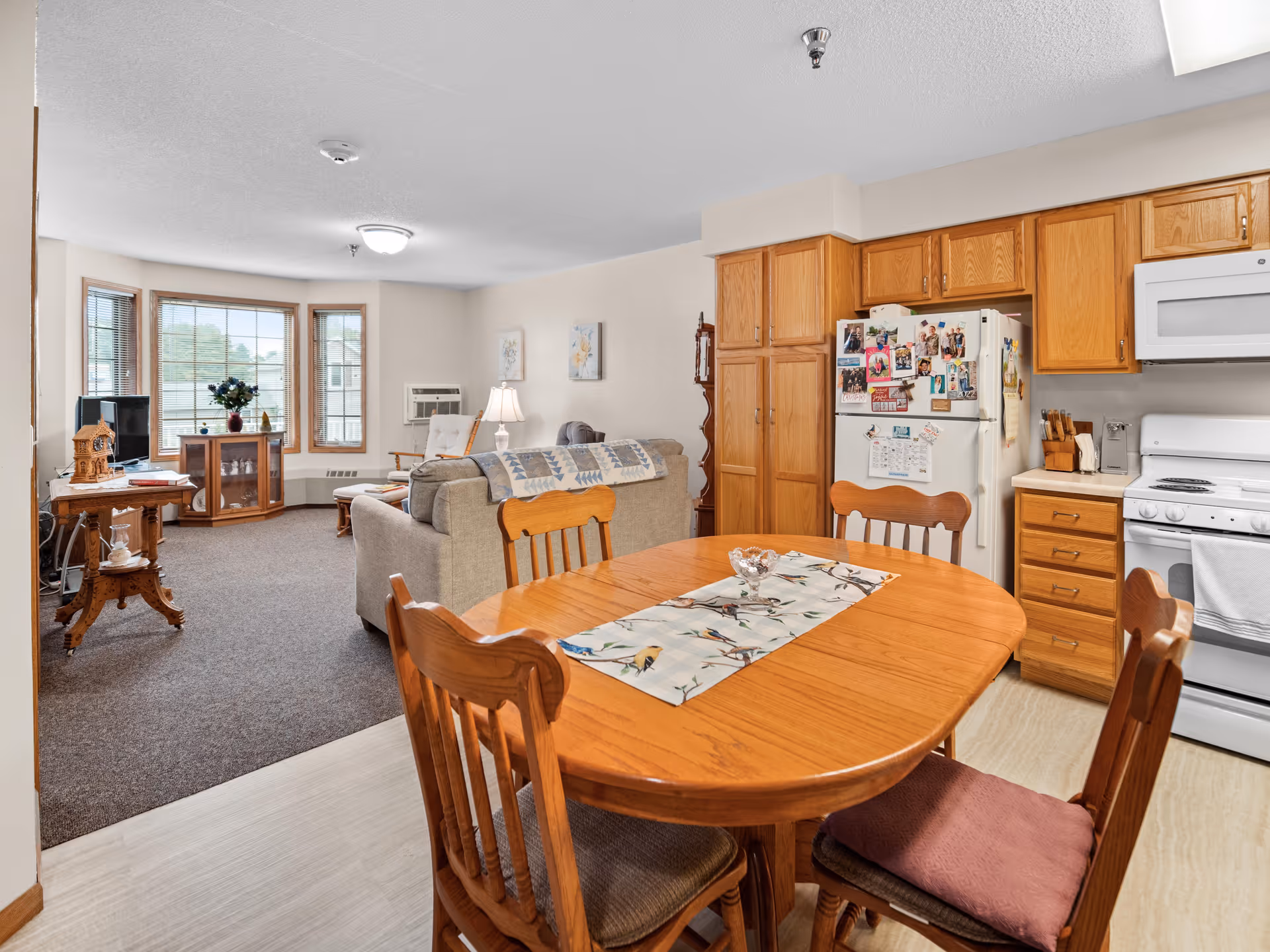 A cozy living and dining area in a senior living facility with a wooden dining table and four chairs in the foreground. The kitchen area features wooden cabinets, a white refrigerator covered with photos and magnets, a white stove, and a microwave. The living room has a beige sofa, a lamp, a TV on a wooden stand, and large windows letting in natural light.