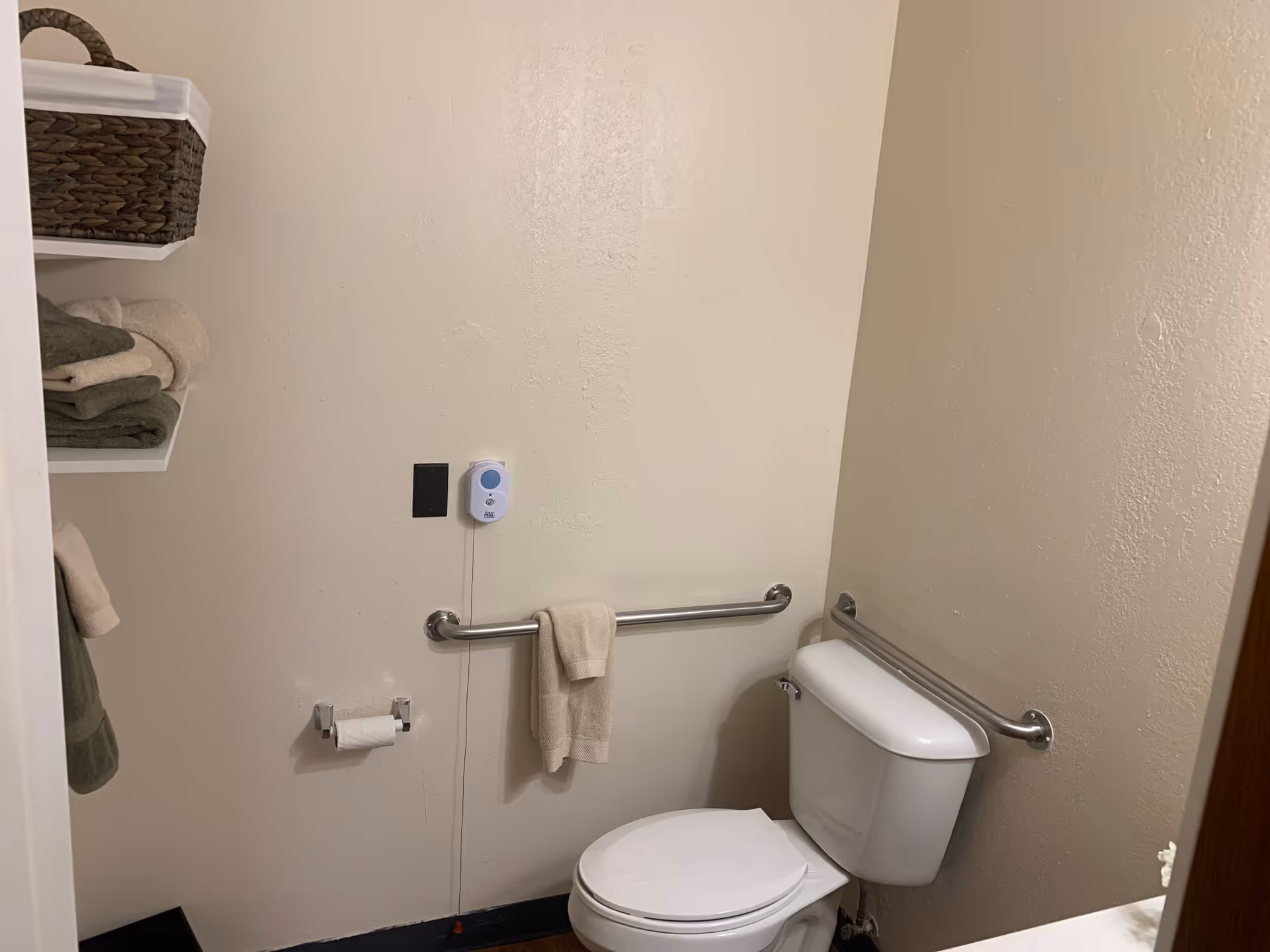 A small bathroom with a white toilet, beige walls, and stainless steel grab bars on two walls. There is a towel hanging on one grab bar and a toilet paper holder with a roll of toilet paper on the wall. Shelves on the left hold folded towels and a woven basket. A white emergency pull cord with a blue button is mounted on the wall above the grab bar.