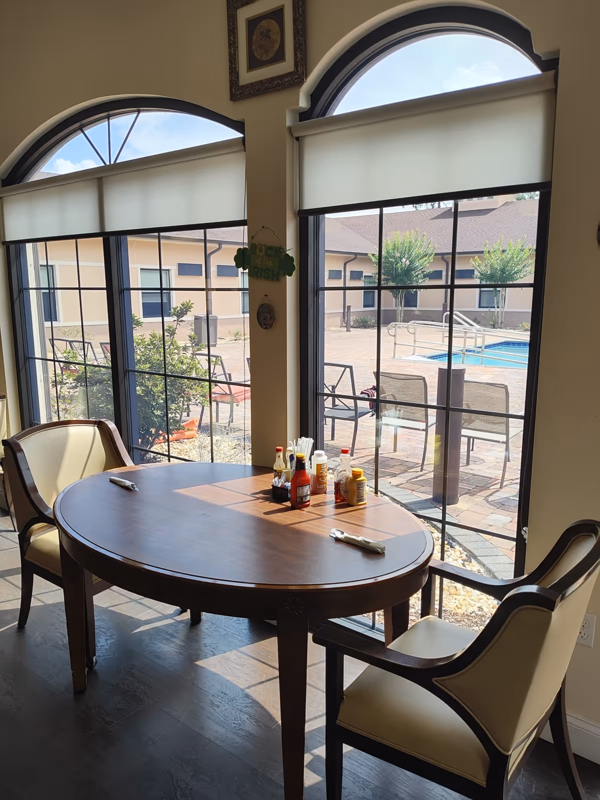 A small dining area with a wooden oval table and two cushioned chairs near large windows with arched tops. On the table are condiments including ketchup, mustard, and salt, along with napkins and utensils. Outside the windows, there is a patio area with chairs and a swimming pool surrounded by a building and some trees.
