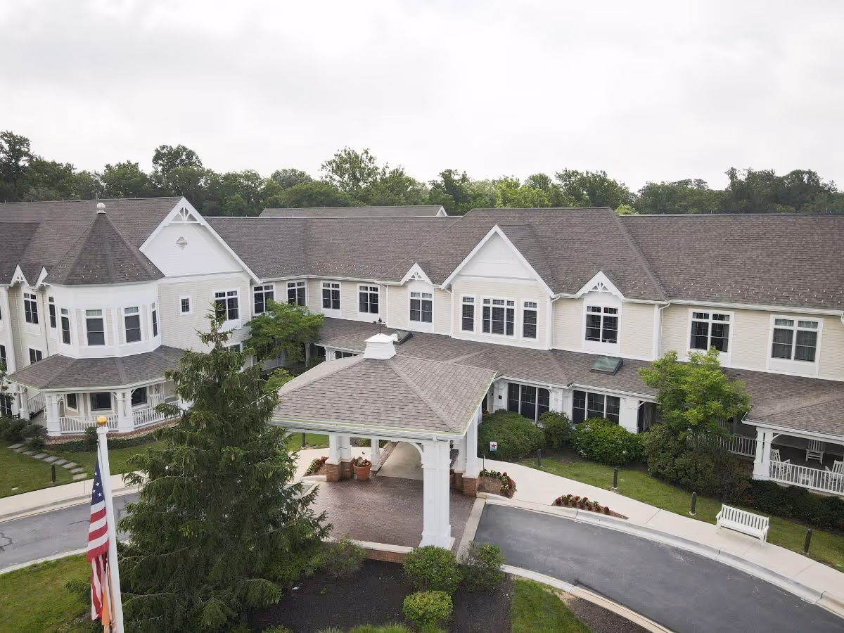 Exterior view of a large, two-story senior living facility building with beige siding and multiple windows. The building has a covered entrance with white columns and a peaked roof. There is a circular driveway in front, landscaped greenery, and an American flag on a flagpole. Trees and a cloudy sky are visible in the background.