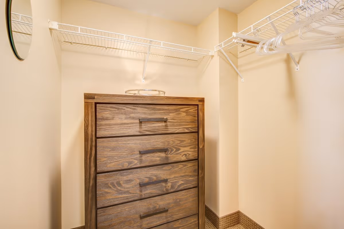 A small walk-in closet with beige walls, white wire shelving on two walls, a wooden chest of drawers with four drawers, and a round mirror partially visible on the left wall.