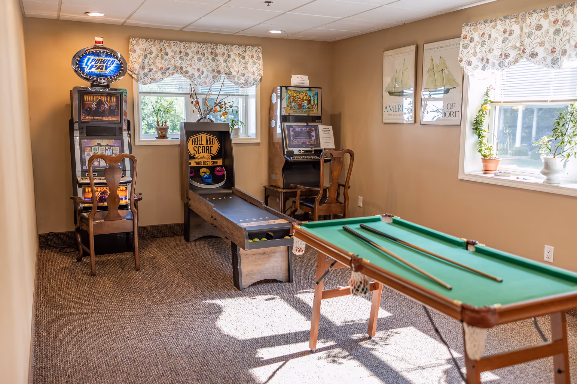 A cozy game room with a small pool table with two cues on it, a skee-ball style game labeled 'Roll and Score', and two vintage slot machines. The room has beige walls, carpeted floor, two windows with floral valances, and framed nautical-themed artwork on the wall. Wooden chairs are placed in front of the slot machines.