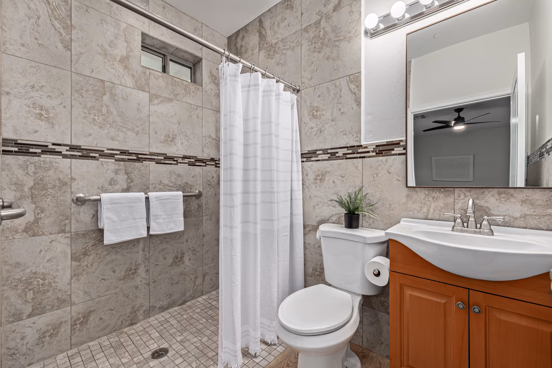 A modern tiled bathroom featuring a shower with a curtain, a toilet, and a wooden vanity with a mirror.