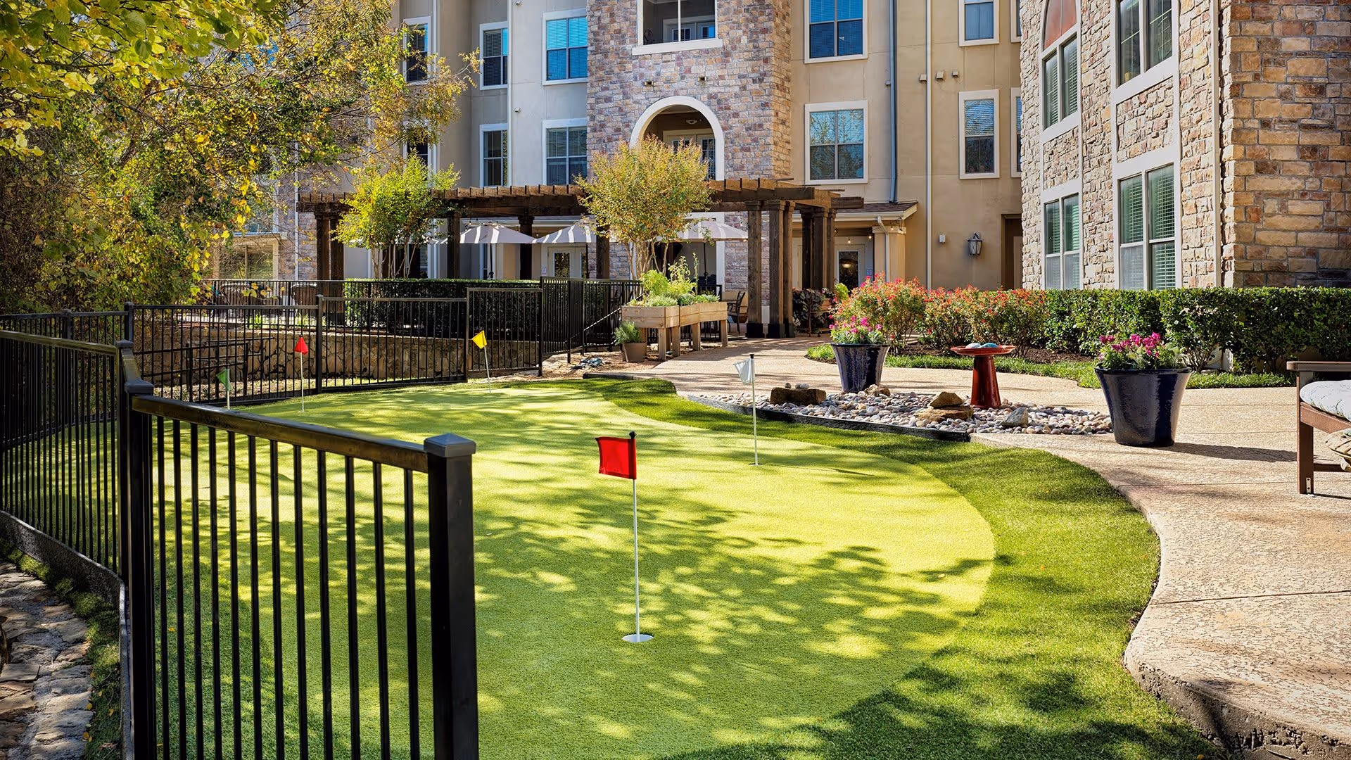 Outdoor putting green with small flags surrounded by a black metal fence, located in a courtyard area of a senior living facility with stone and stucco buildings, potted plants, and a shaded pergola in the background.
