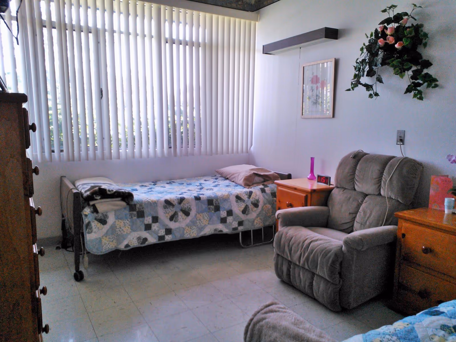 A small bedroom in an assisted living facility with a single bed covered by a patchwork quilt, a cushioned armchair, two wooden nightstands, a framed floral picture on the wall, and a hanging plant. Vertical blinds cover the window, allowing natural light to enter the room.