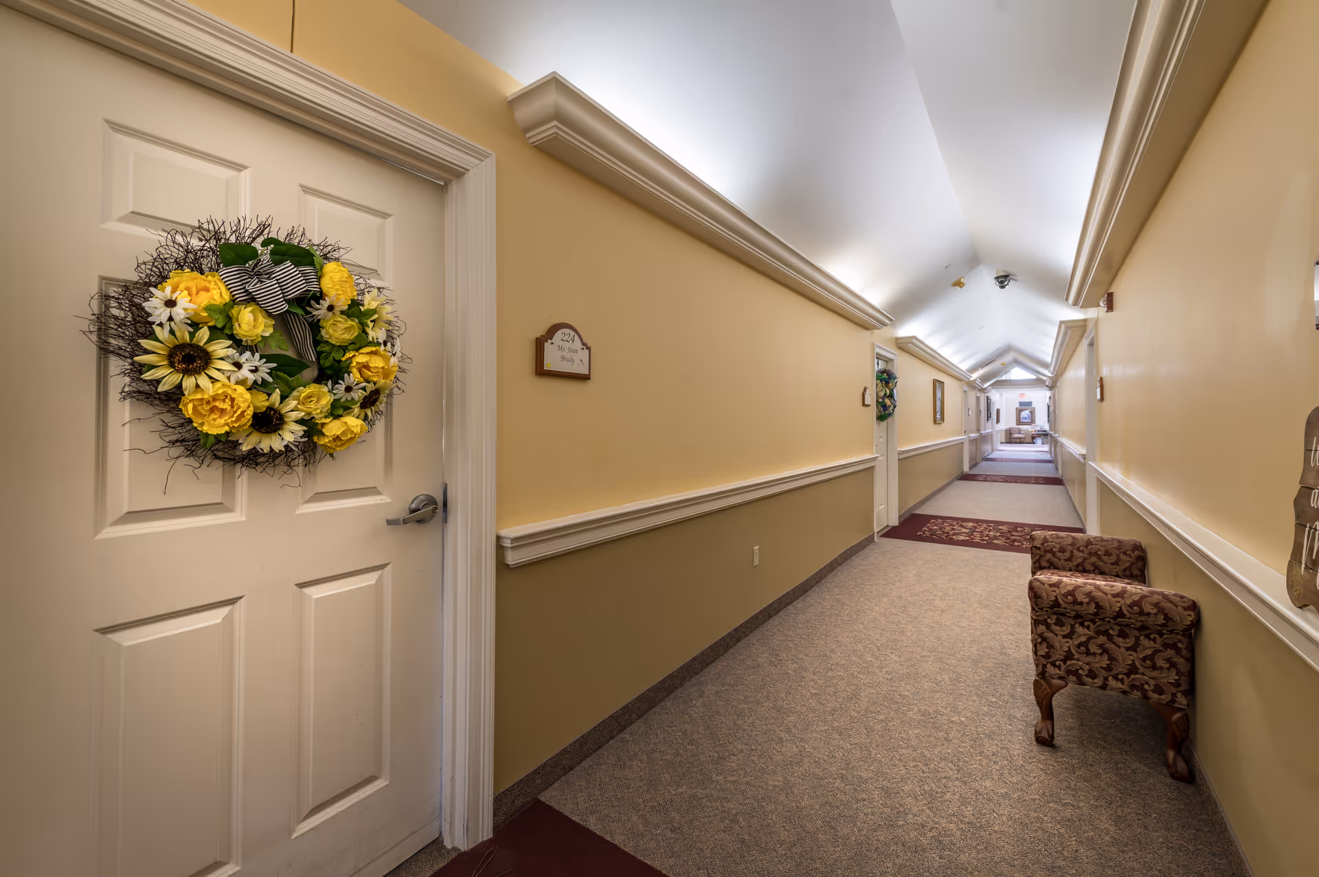 A long, well-lit hallway in an assisted living facility with beige walls and carpeted floor. Doors line the hallway, each decorated with a floral wreath. A patterned armchair is placed along the right wall, and the ceiling has recessed lighting.
