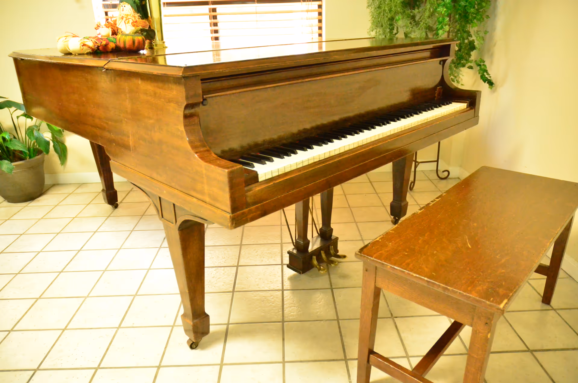 A wooden grand piano with a matching bench on a tiled floor in a bright room with potted plants.