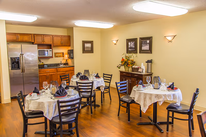 A dining room with three round tables covered with white tablecloths, each set with plates, cups, wine glasses, and black napkins. The room has wooden flooring, beige walls, and wooden cabinets with a stainless steel refrigerator and microwave. There are framed pictures on the walls and a sideboard with a flower arrangement and a coffee dispenser.