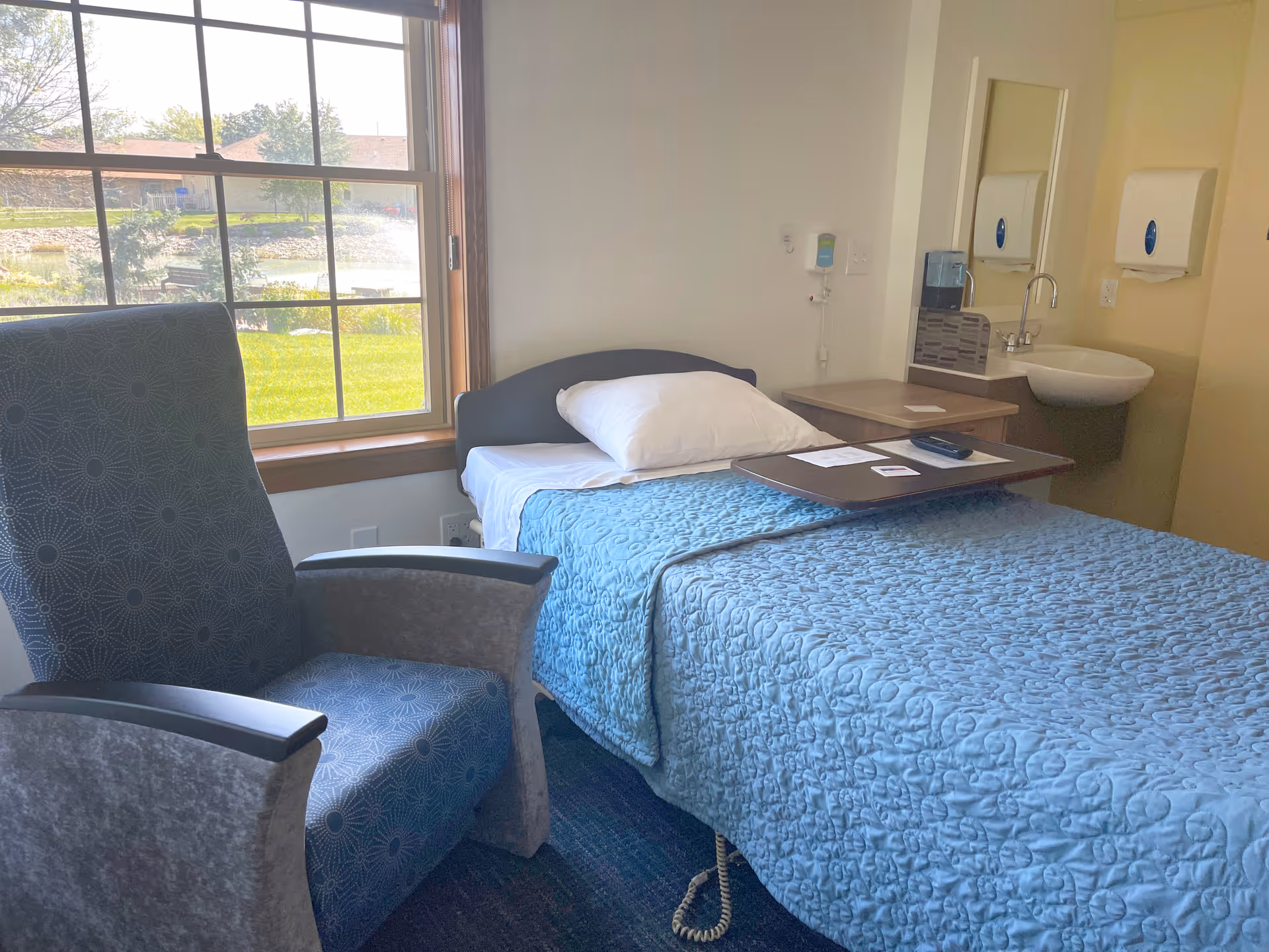 Patient room with a single bed covered by a blue quilt, an overbed tray, a reclining chair, a sink and a window showing grass and a pond.