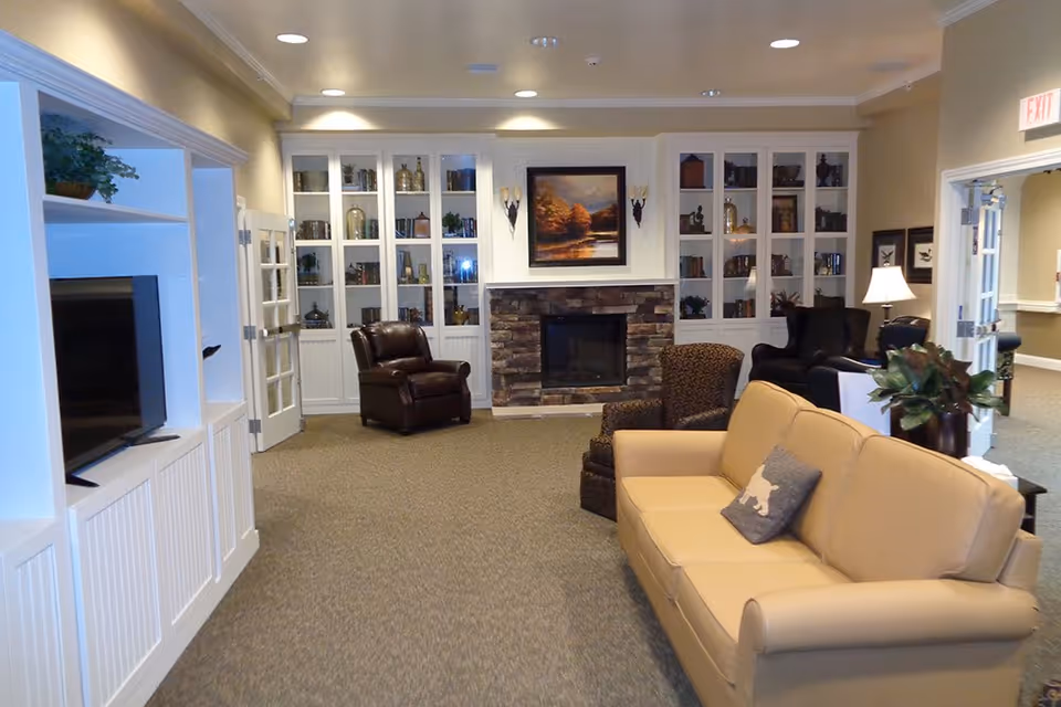 A cozy living room area in a senior living facility featuring a beige sofa with a decorative pillow, several armchairs, a stone fireplace with a framed landscape painting above it, built-in white cabinets with glass doors displaying decorative items and books, a flat-screen TV on the left, and soft lighting from ceiling fixtures and a table lamp.