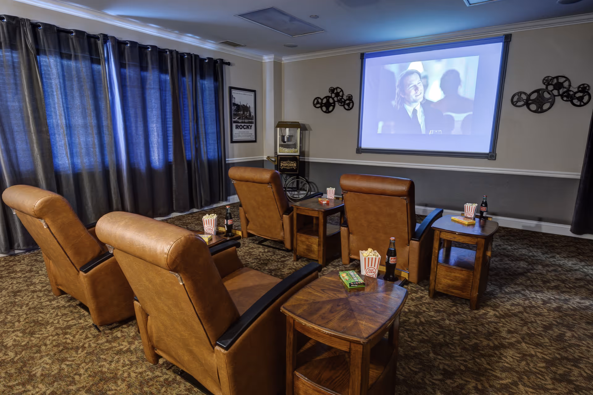 A cozy movie theater room with four brown leather recliners arranged in two rows facing a large projection screen. Each recliner has a small wooden side table with popcorn, soda bottles, and snacks. The room has dark curtains covering the windows, a popcorn machine in the corner, and movie-themed wall decorations.