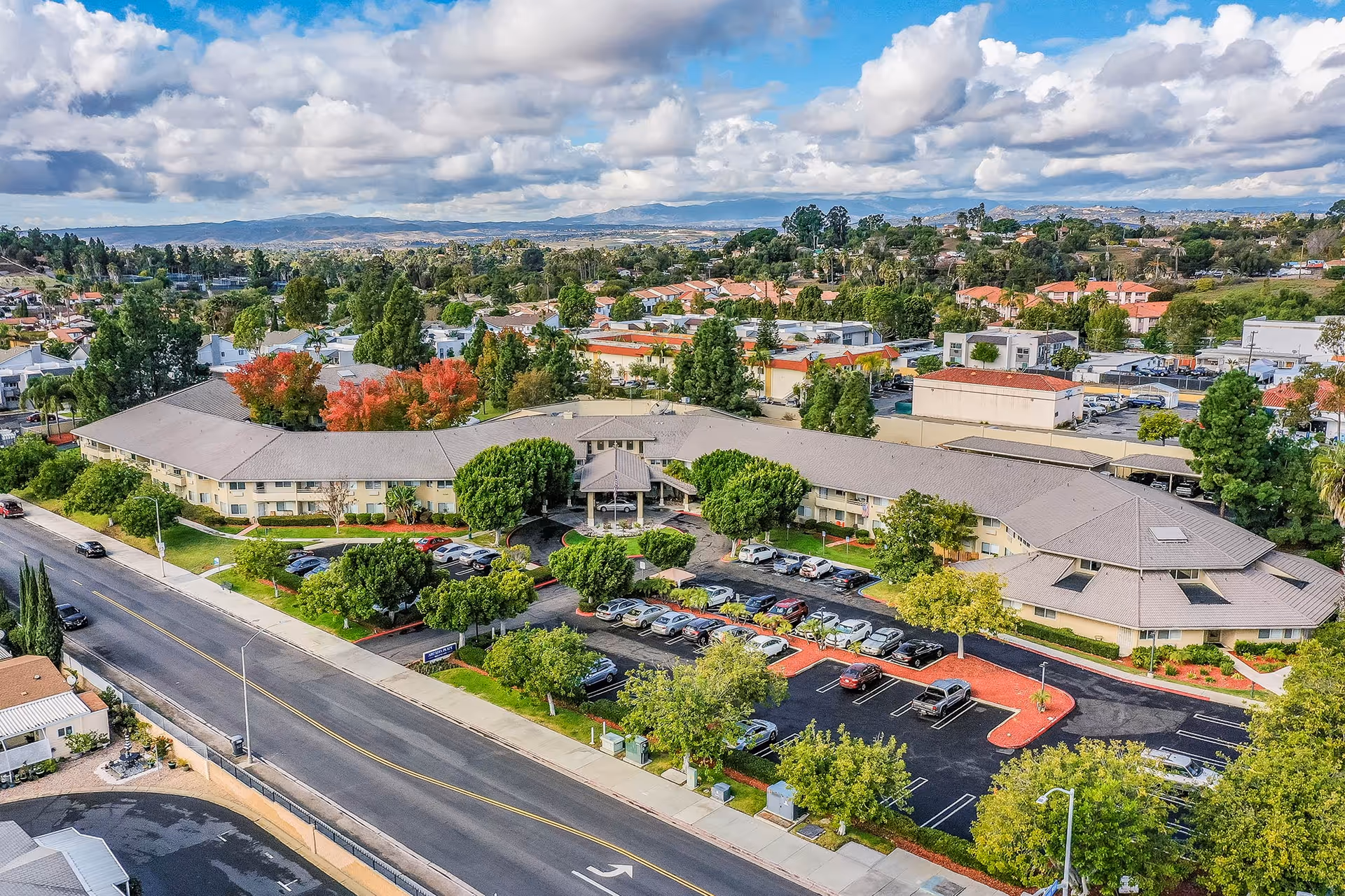 Aerial view of Arcadia Place Senior Living facility showing a large, U-shaped building surrounded by trees and greenery. The parking lot in front of the building has several cars parked. The surrounding neighborhood and distant hills are visible under a partly cloudy sky.
