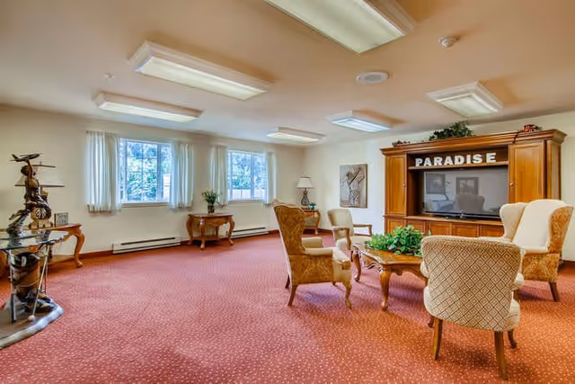 A spacious living room with red carpet and cream-colored walls, featuring a wooden entertainment center with the word 'PARADISE' on top, a flat-screen TV, four upholstered chairs arranged around a wooden coffee table with a green plant centerpiece, two windows with white curtains letting in natural light, a side table with a lamp, and decorative sculptures on the left side.