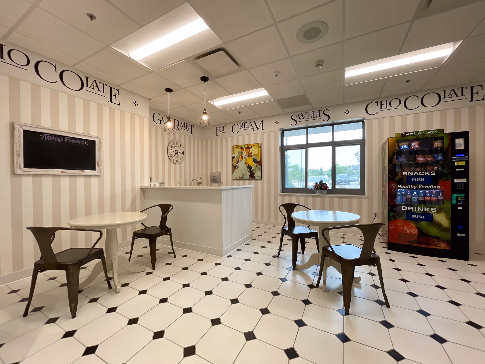 A small dining area with two round white tables and four metal chairs. The walls have vertical beige and white stripes with decorative text near the ceiling reading 'CHOCOLATE GOURMET ICE CREAM SWEETS'. There is a vending machine labeled 'SNACKS' and 'DRINKS' on the right side, a window with a view outside, and a counter with a clock and a small chalkboard on the wall.