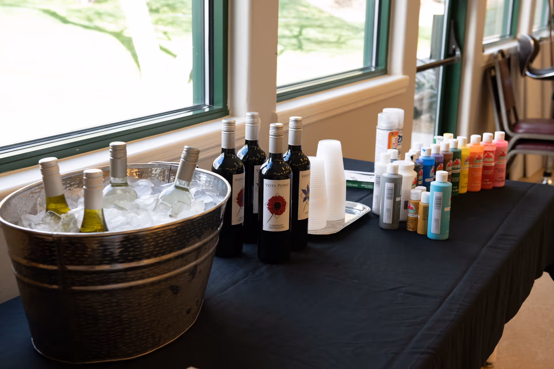 A table covered with a black cloth displaying bottles of white and red wine, with the white wine bottles chilling in a metal ice bucket. Next to the wine bottles are clear plastic cups and a variety of colorful paint bottles arranged in a row. The table is set up near large windows with a view of greenery outside.