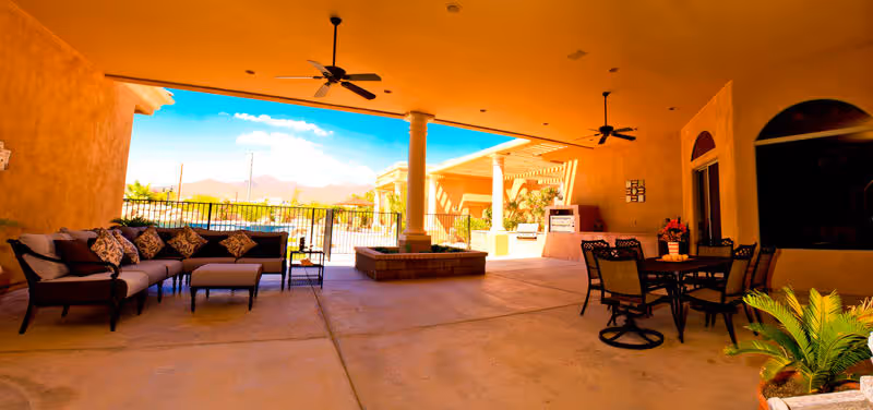Covered outdoor patio area with ceiling fans, a seating area with cushioned sofas and pillows on the left, a dining table with chairs on the right, potted plants, and a view of a fenced pool and mountains in the background under a bright blue sky.