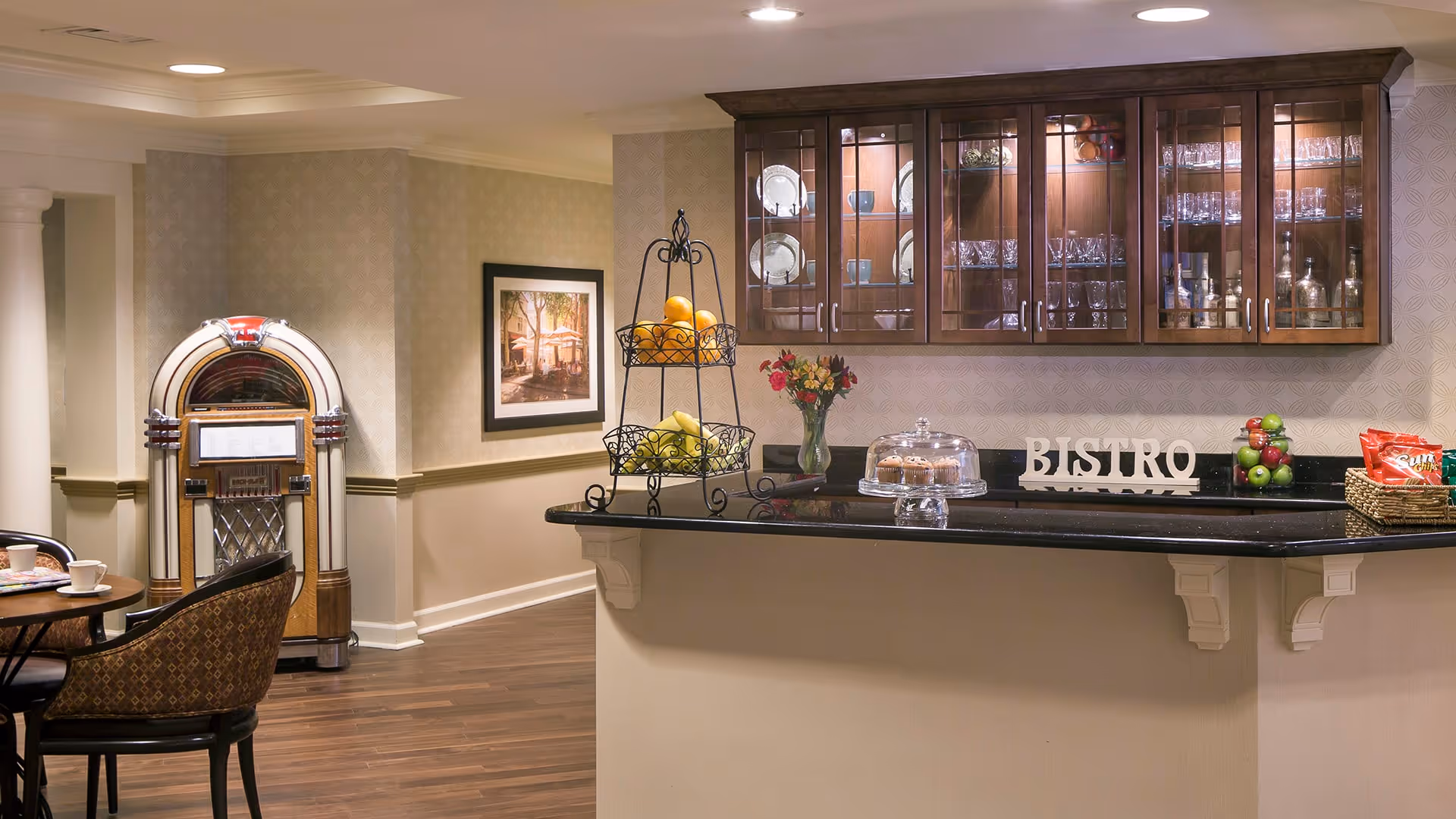 Interior view of a bistro area in a senior living facility featuring a counter with a black countertop, a tiered fruit basket, a vase with flowers, a glass cake stand with muffins, and a decorative 'BISTRO' sign. Behind the counter are wooden cabinets with glass doors displaying plates, cups, and glassware. To the left, there is a vintage jukebox, a framed picture on the wall, and a small round table with chairs.
