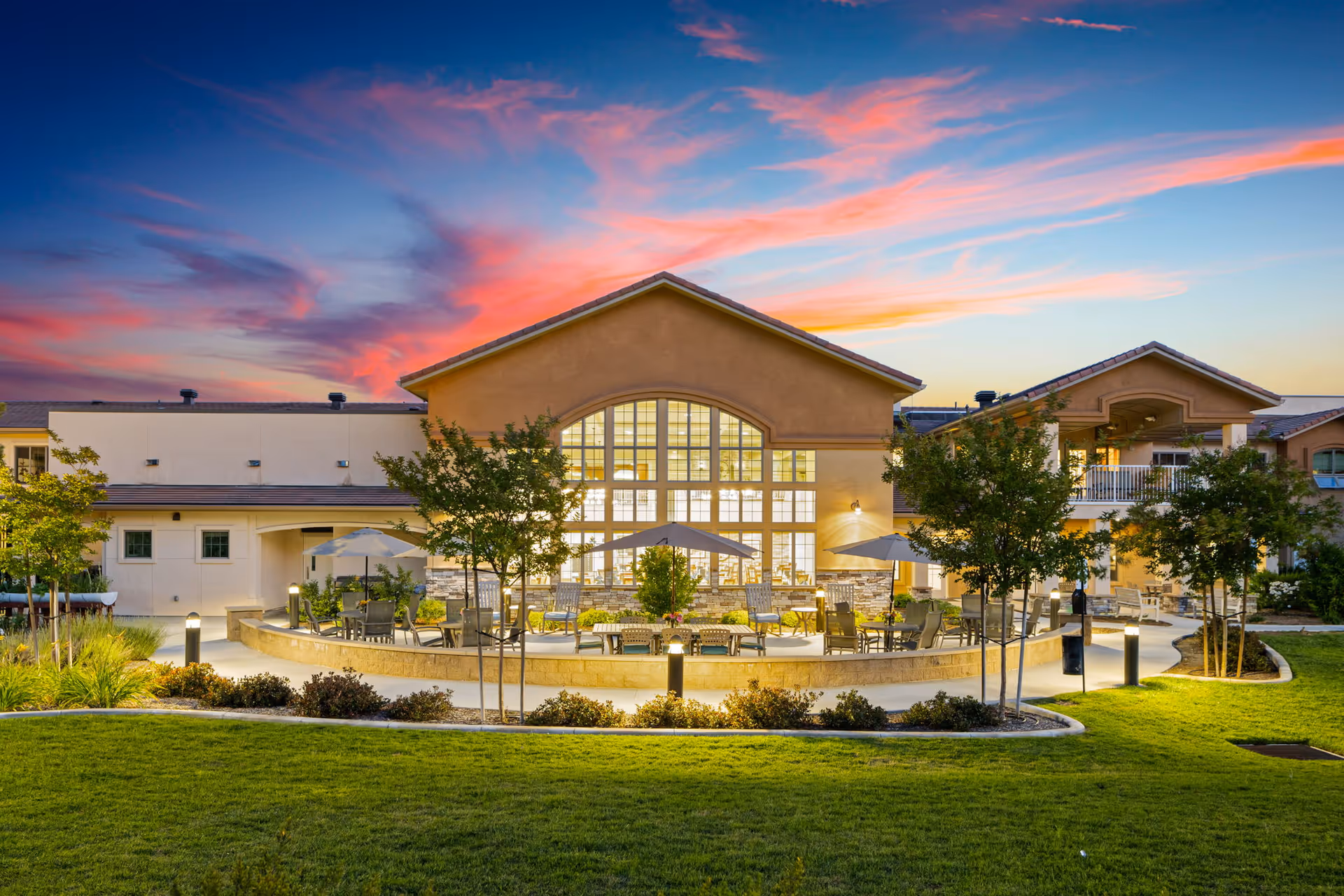 Exterior view of Ivy Park At Murrieta senior living facility at sunset, showing a well-lit building with large windows, outdoor seating areas with umbrellas, trees, and a manicured lawn under a colorful sky.