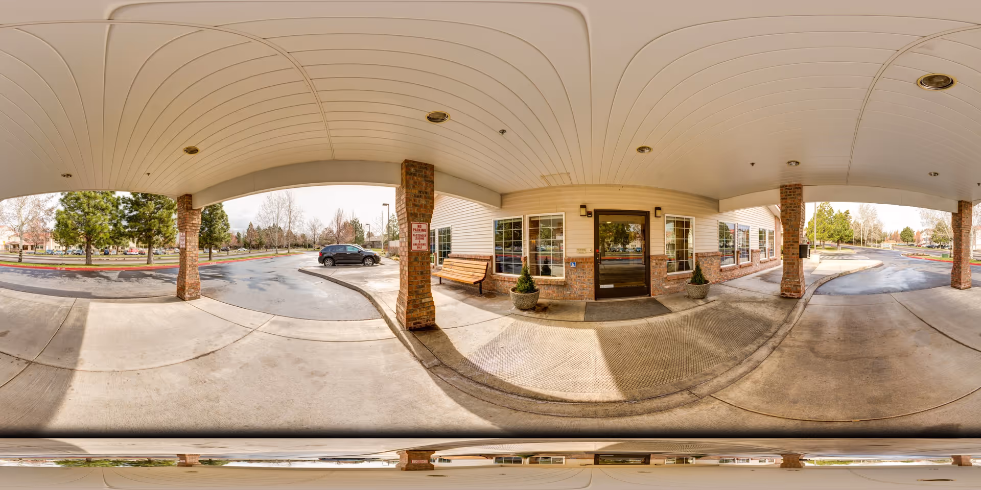 Covered entrance area of Aspen Ridge Memory Care facility with brick pillars, a bench, potted plants, and a glass door leading inside. The driveway and parking area are visible with some trees and a parked car in the background.