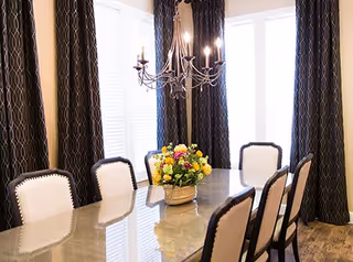 A dining room with a long rectangular table surrounded by eight white upholstered chairs with dark trim. A floral centerpiece sits on the table. The room has large windows covered with dark patterned curtains and a chandelier with lit candles hanging above the table.