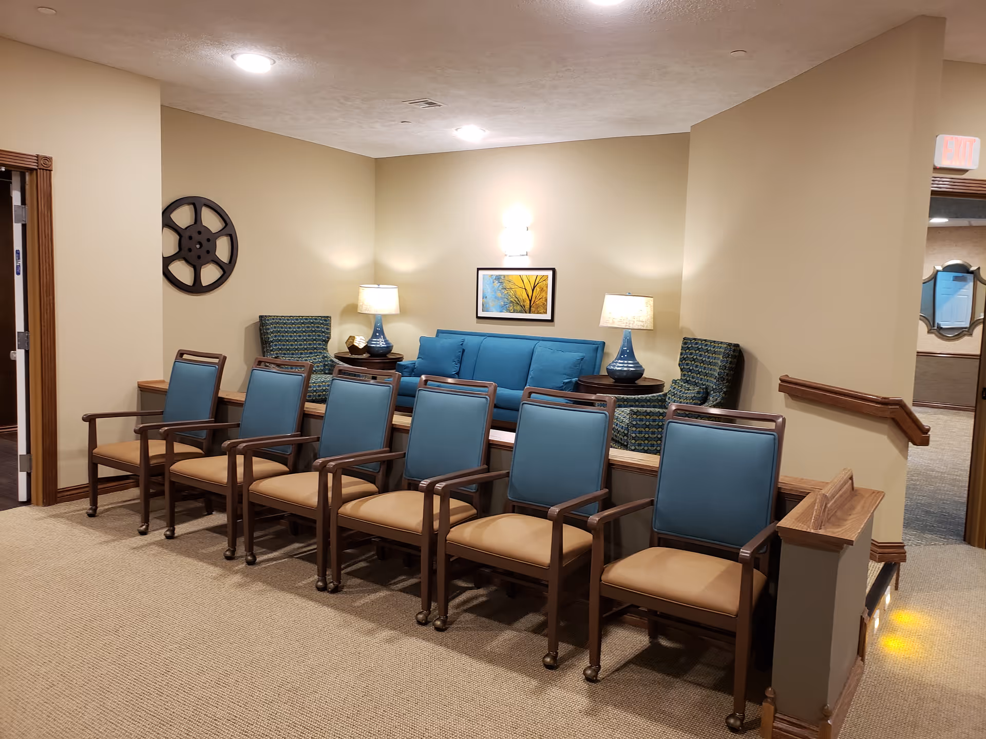 A seating area in a retirement community featuring a row of six wooden chairs with blue cushions and beige seats. Behind the chairs is a blue sofa flanked by two patterned armchairs and two table lamps with blue bases. The walls are beige with a framed picture and a decorative film reel mounted on the wall. The space is well-lit with ceiling lights and wall sconces.