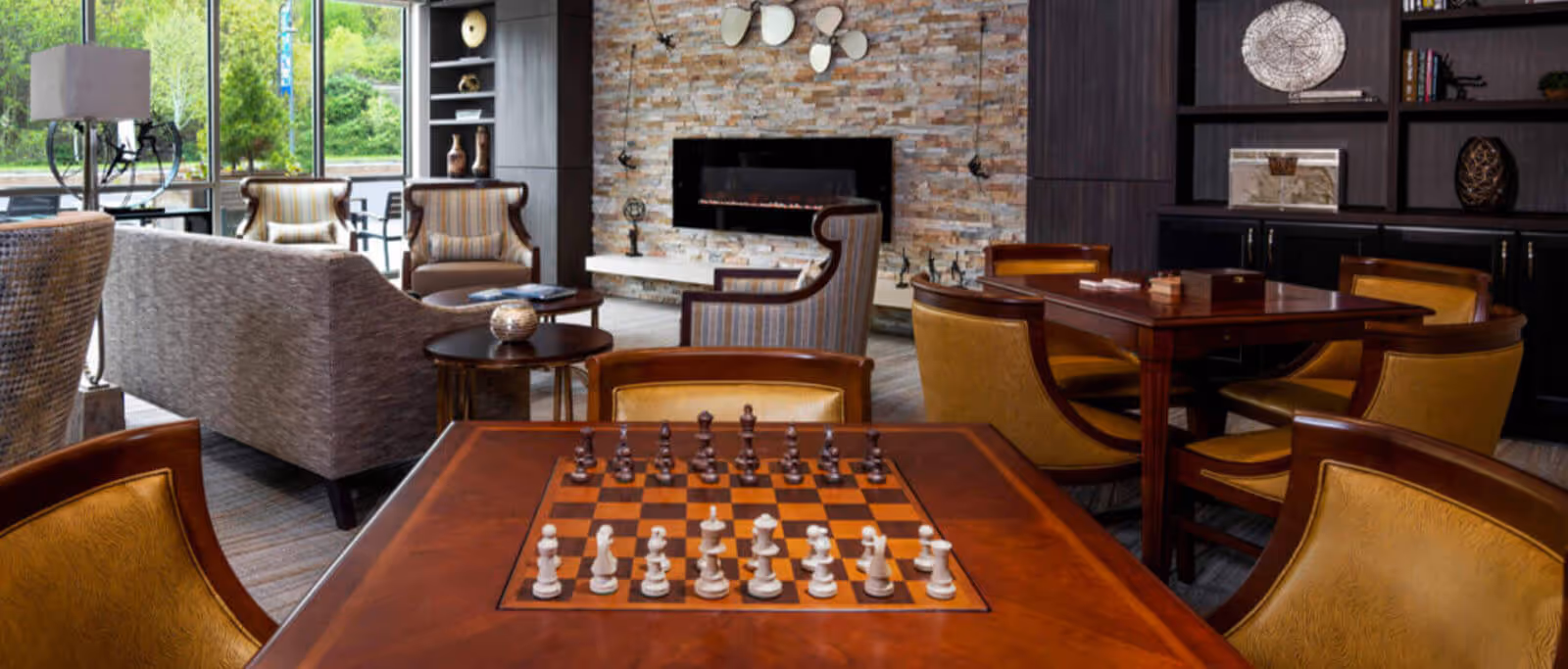 A cozy common area in a senior living facility featuring a wooden chess table with chess pieces set up, comfortable upholstered chairs, a modern electric fireplace set in a stone accent wall, large windows letting in natural light, and built-in dark wood shelves with decorative items.