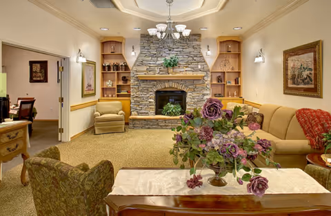 A cozy living room with a stone fireplace in the center, flanked by built-in wooden shelves. There is a beige sofa with a red throw blanket on the right, an armchair on the left, and a floral arrangement on a wooden table in the foreground. The room has warm lighting and framed artwork on the walls.