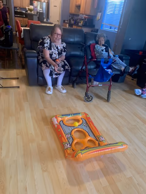Two elderly women sitting on a dark gray leather couch in a living room with light wooden flooring. One woman is wearing a floral dress and white slippers, smiling and reaching towards an inflatable orange game on the floor. The other woman, seated next to a red walker, is wrapped in a blanket. The background shows a kitchen area with wooden cabinets and a window with blinds.