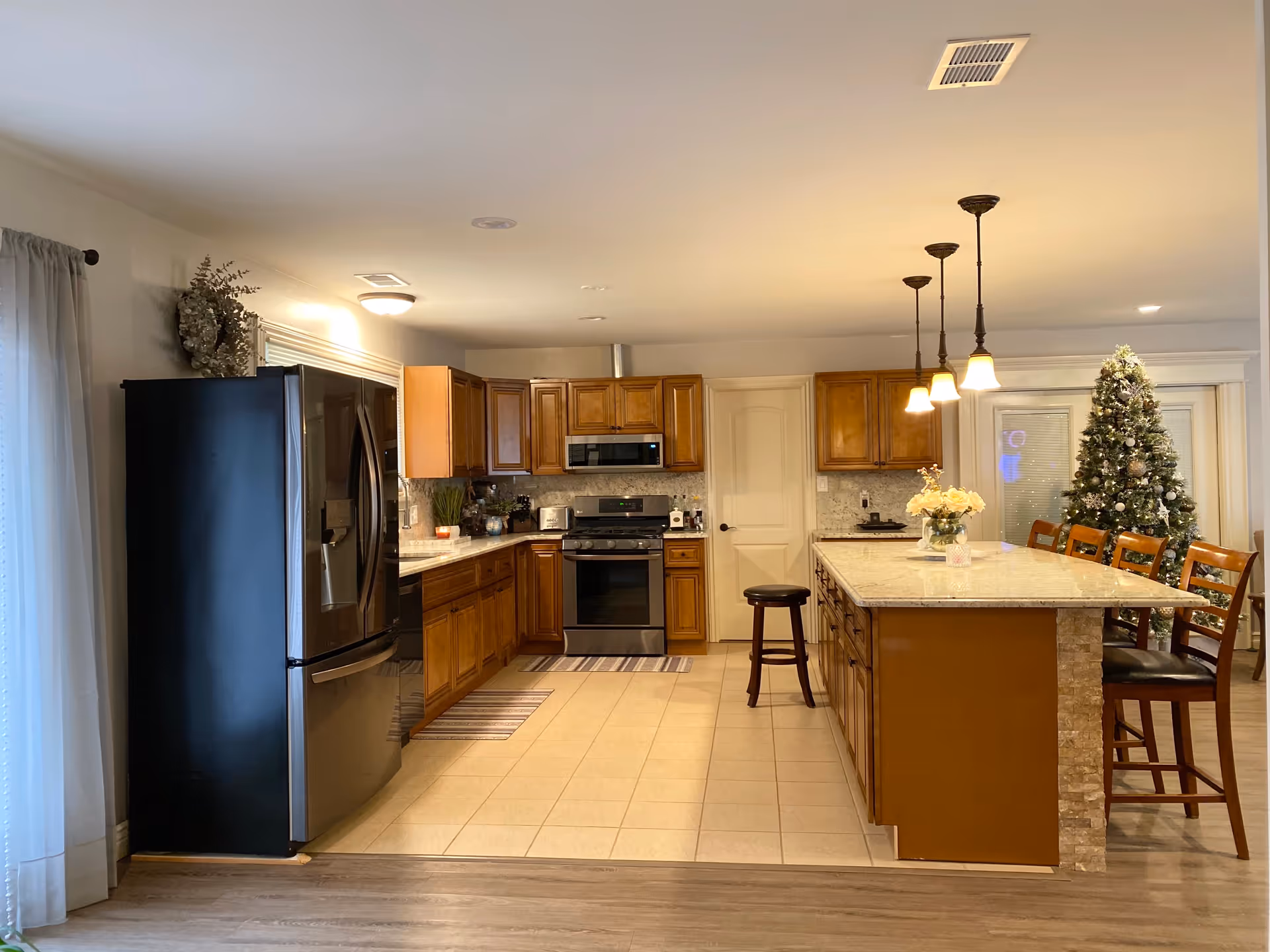 A spacious kitchen with wooden cabinets, a black refrigerator, a stove with a microwave above it, and a large island with a granite countertop. The island has several wooden chairs and a vase with flowers on top. Three pendant lights hang above the island. In the background, there is a decorated Christmas tree near a glass door.